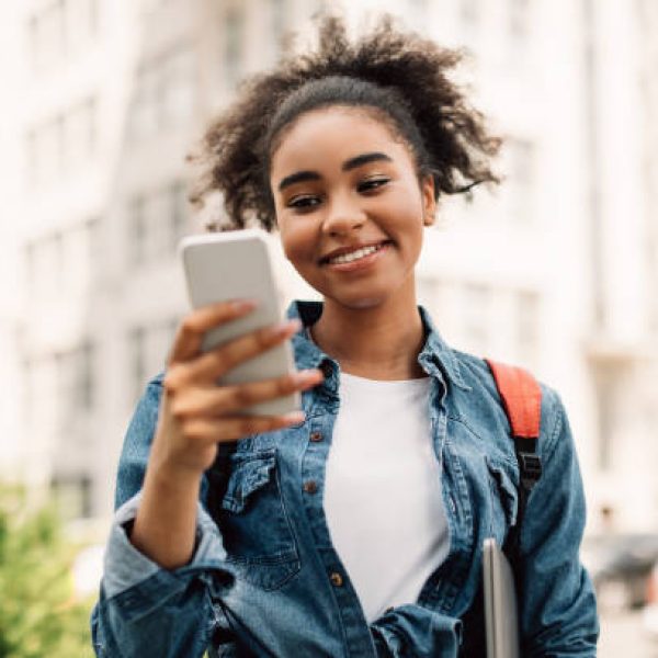 Cheerful African American Student Girl Using Mobile Phone Texting And Learning In Educational Application Standing Outside Near College Building. New App For Your Cellphone Concept